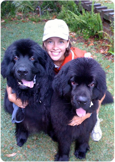 A smiling woman in a cap hugging two large, fluffy black Newfoundland dogs on a grassy lawn