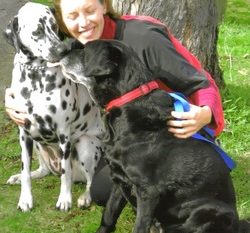 A smiling woman embracing a black dog and a Dalmatian in a park setting.